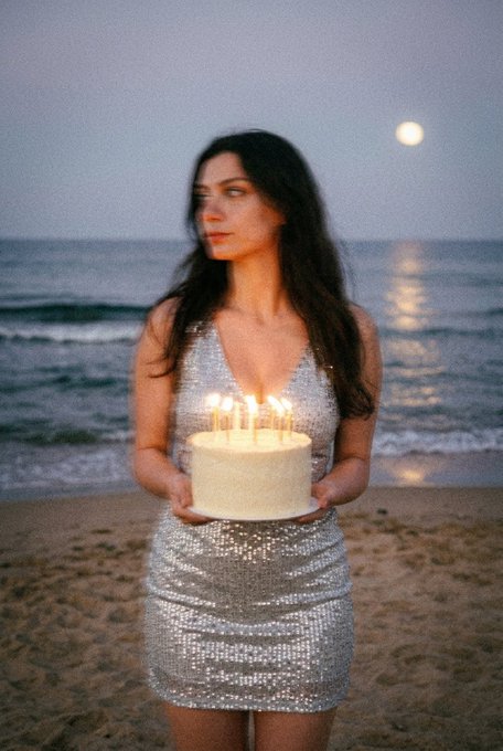 The image features a woman standing on a beach holding a white cake with lit candles. The background of the photo shows an ocean view at night, creating a serene atmosphere for her birthday celebration. The caption "Late Gm frens Today is my birthday! I’m not getting older, I’m just becoming a classic" suggests that she is embracing her age and enjoying life's simple pleasures like spending time on the beach with friends or family while celebrating her special day.