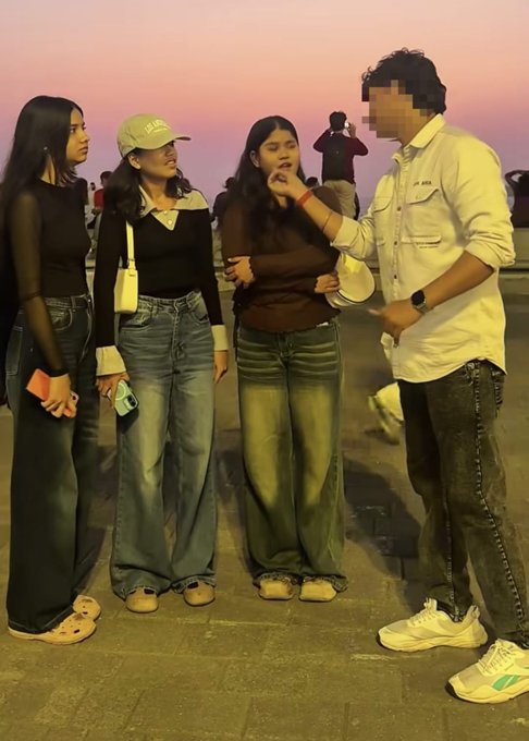 A group of four young people stand together at sunset near a coastline, with three women in casual outfits including jeans and sweaters, and a man in a white shirt whose face is blurred.