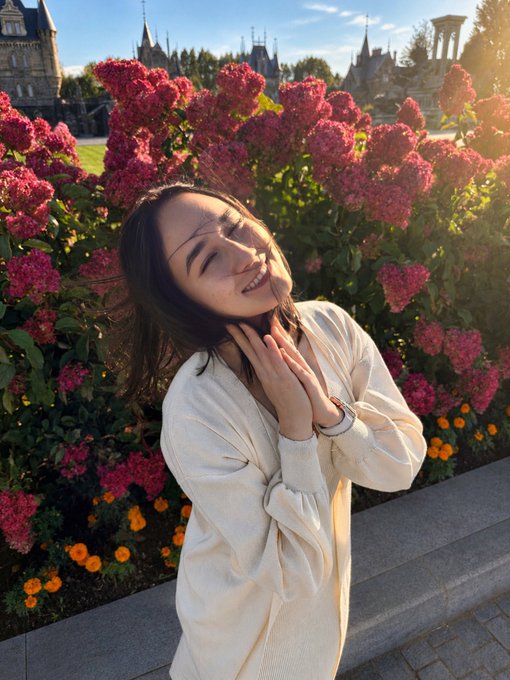 A woman in a cream-colored sweater smiles with her eyes closed, standing in front of vibrant pink and orange flowers with a historic castle in the background.