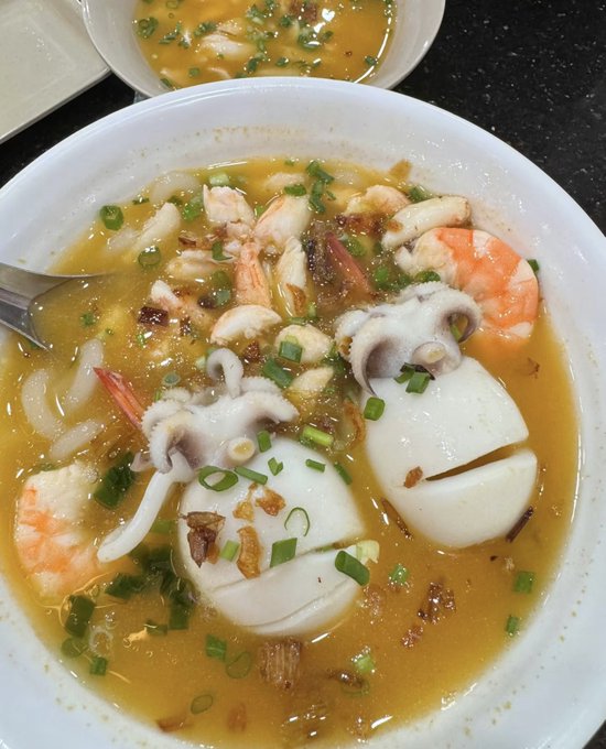 A bowl of seafood soup featuring shrimp, octopus, and tofu in a savory broth, garnished with green onions and fried garlic. The image contrasts with the post's playful mention of choosing instant noodles versus a more substantial meal, showcasing a hearty lunch option that's far more extravagant than the budget-friendly ramen referenced in the message.