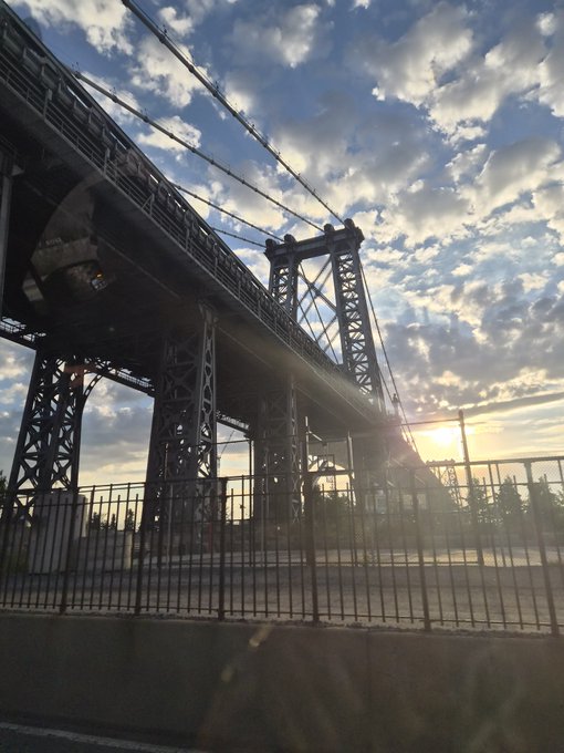 An angled view of a large steel truss bridge silhouetted against a partly cloudy sky at sunset, with a metal fence in the foreground.