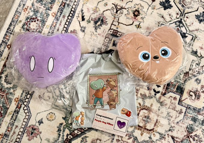 Two plush toys in plastic packaging sit on a patterned rug; one is purple and the other brown, next to a printed certificate that reads 'Congratulations! You won a Purple Hug Pillow'.