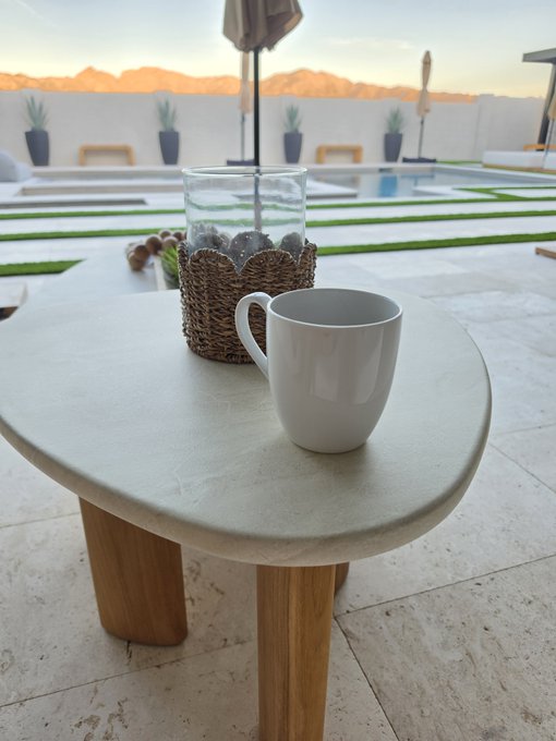 A white ceramic coffee mug sits on a round stone table beside a woven candle holder, with a pool, potted plants, and mountains visible in the background during sunset.