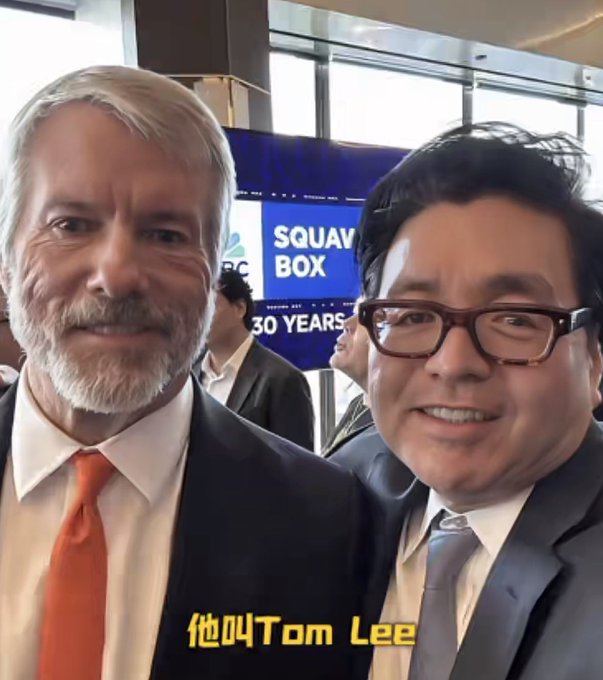Two men in suits pose for a selfie, one older man with white hair and an orange tie, the other wearing black glasses, with a 'SQUAWK BOX' sign in the background and yellow Chinese text '他叫Tom Lee' at the bottom.