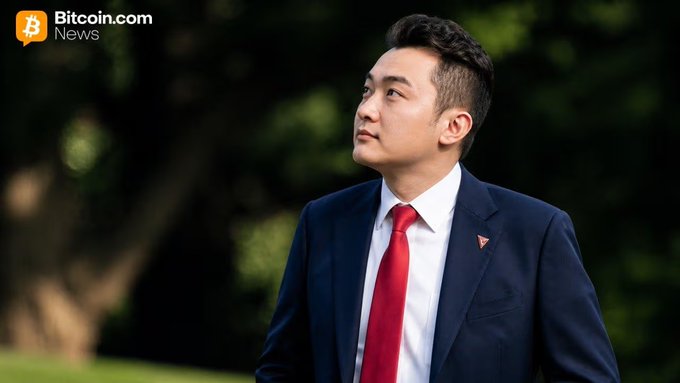 A Bitcoin.com News photo of an Asian man in a dark suit and red tie, looking off to the side in an outdoor setting.