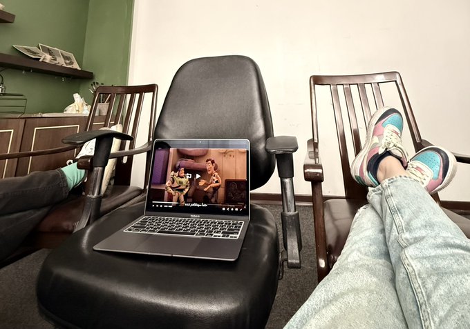 A first-person view of someone with their feet up wearing colorful sneakers, watching a video on a MacBook Pro placed on an office chair in what appears to be a meeting room.