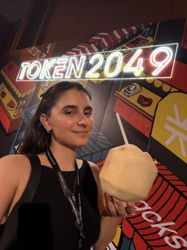 A selfie of a smiling woman in a black top holding a fresh coconut, standing in front of a colorful mural with a neon sign reading 'TOKEN2049'.