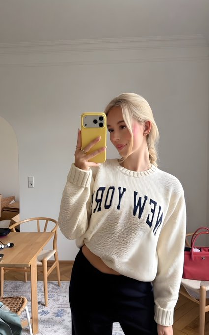 A blonde woman takes a mirror selfie wearing a cream sweater reading "NEW YORK" while asking her followers for "GM" (good morning) greetings. The casual morning setup with a yellow phone case and bright room complements her friendly request for morning interactions.