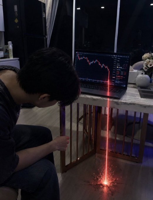 Dark humorous photo of a person with head in hands looking at a laptop showing a steep red downward stock/crypto chart, with a glowing red laser beam metaphorically "burning a hole" through the floor.