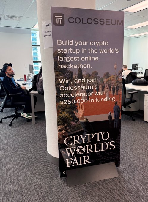 A 'Crypto World's Fair' promotional banner from Colosseum advertising a crypto hackathon with $250,000 in accelerator funding, displayed in an office setting.