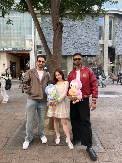A photo of two men and a woman posing on a city sidewalk, where the woman in the center is holding two colorful stuffed animal plushies.