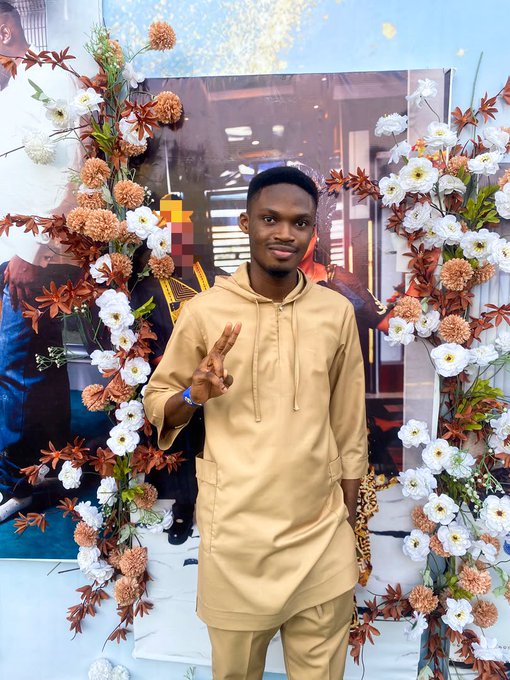 A young man wearing a tan traditional tunic outfit standing in front of a floral archway decorated with white and orange flowers, making a peace sign.