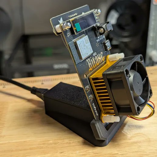 The image shows a Bitaxe Bitcoin mining device mounted on a black stand with a cooling fan attached, prominently displaying "Bitaxe" branding on its circuit board along with visible components like a chip module and power connector. This perfectly illustrates the post's message about upgrading from a loose, Wi-Fi-only setup to a tidier configuration with the included stand, showcasing the cleaner mining arrangement being promoted.