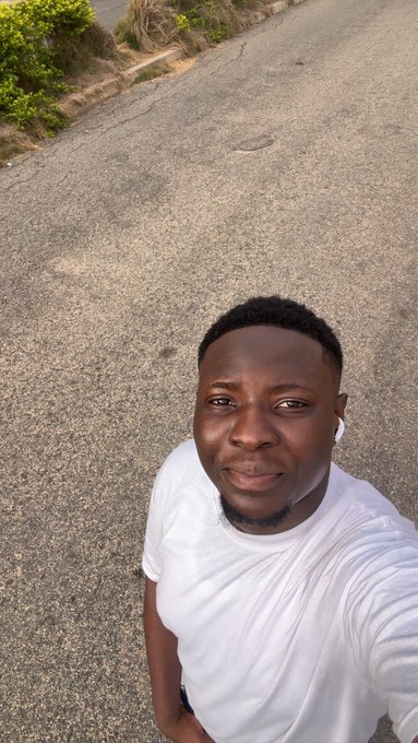 A young man wearing a white t-shirt and wireless earbuds takes a selfie while standing on an empty road, casually introducing himself with a relaxed expression. The laid-back personal vibe complements his friendly Tuesday greeting, matching the approachable tone of sharing crypto news about Emblem ($Emblem) wrapping Bitcoin Ordinals.
