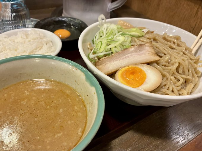 A Japanese meal set featuring a bowl of tsukemen-style dipping ramen with chashu pork and a soft-boiled egg, a side of white rice, a raw egg, and a bowl of dipping broth.