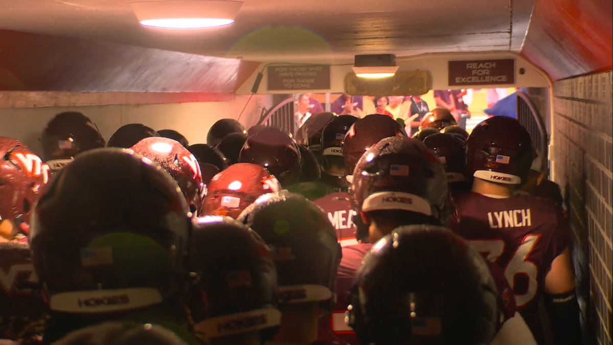 Virginia Tech football players don maroon "Hokies" helmets as they crowd through a tunnel toward the field, with motivational signs reading "FOR THOSE WHO HAVE PASSED FOR THOSE TO COME" and "REACH FOR EXCELLENCE" overhead, capturing the team's pregame entrance as the crowd welcomes them with Enter Sandman ahead of their primetime matchup against Pitt.