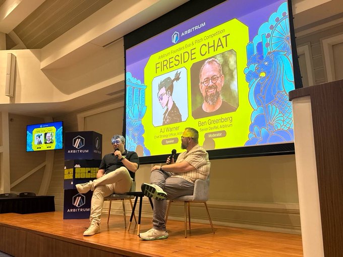 Two men, identified on screen as AJ Warner (Chief Solutions Officer, Arbitrum) and Ben Greenberg (Senior DeFi Lead, Arbitrum), are seated on stage holding microphones during a "Fireside Chat" at the Arbitrum Founders Eve & Pitch Competition in Singapore, with their photos and social handles (@ajwarner90 and @hummusonrails) displayed on the large presentation screen behind them.