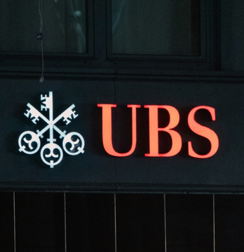 A close-up photograph of the UBS logo illuminated in orange/red against a dark building exterior, featuring the company's signature crossed keys emblem with three circles.