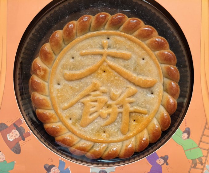 A large, decorative Chinese pastry or bread is displayed on a black tray, featuring the characters '大蘇' (Da Su) embossed in the center surrounded by a twisted bread border, sitting on an orange surface with cartoon figures.