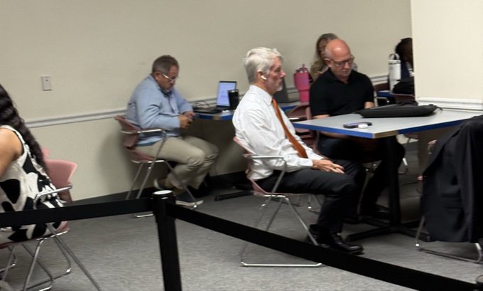 A group of people seated in what appears to be a meeting or classroom setting, with a man in a white shirt and orange tie in the foreground and others working on laptops in the background.