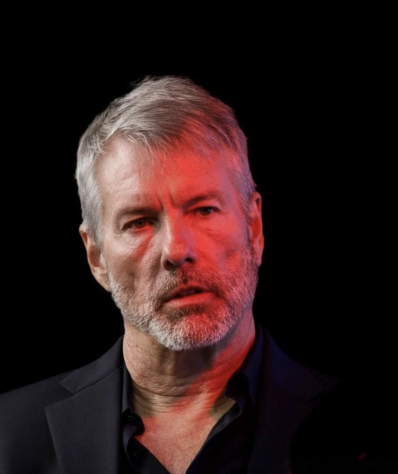 A close-up portrait of a gray-haired, bearded man in a dark suit and black shirt, dramatically lit against a black background with red lighting accents on his face.