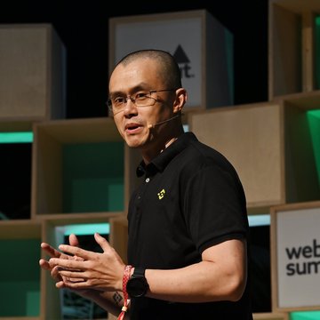 A man wearing a black polo shirt and glasses speaks on stage at what appears to be a Web Summit event, gesturing with his hands.