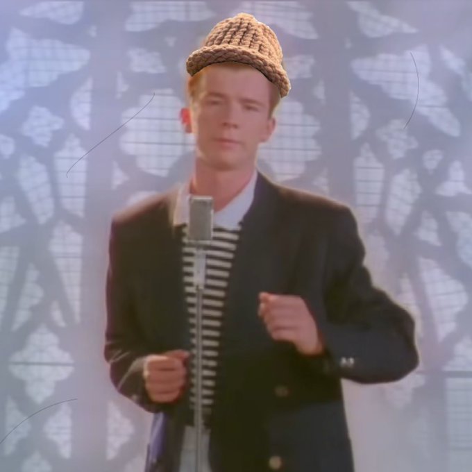A young man with blonde hair wearing a woven tan hat, black and white striped shirt, and dark blazer stands in front of a microphone against a patterned backdrop, with his fist raised mid-performance.
