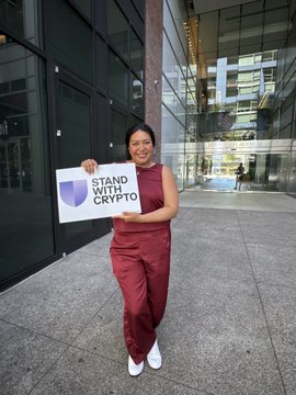 A woman in a burgundy jumpsuit stands outside a modern building holding a sign that reads 'STAND WITH CRYPTO'.