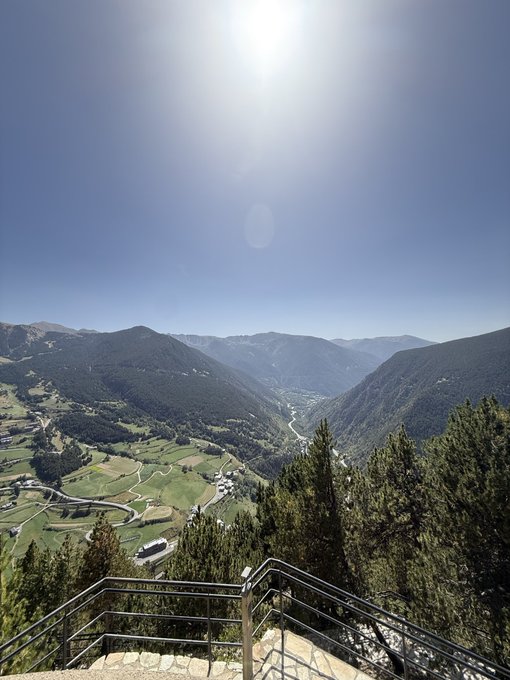 A scenic mountain landscape view from an elevated lookout point with metal railings, overlooking a green valley with small villages, winding roads, and forested mountains under a bright blue sky.