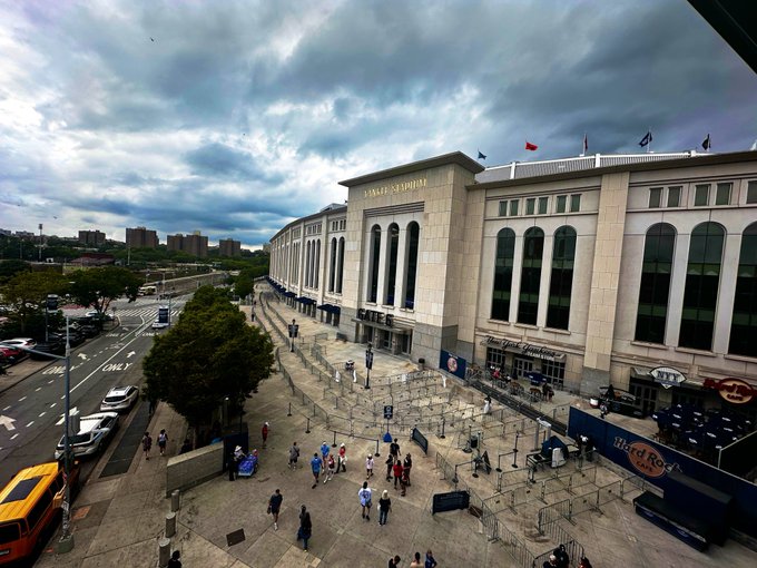 An aerial view of Yankee Stadium in New York, showing the grand entrance with pedestrians walking on the plaza, under a cloudy sky with a yellow taxi visible on the street.