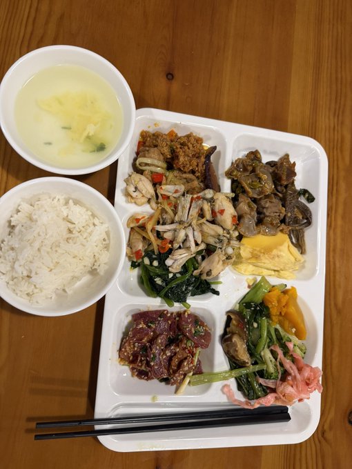 A Korean-style meal featuring a white tray with multiple compartments containing various banchan (side dishes) like beef, vegetables, and pickled radish, accompanied by a bowl of white rice and a bowl of clear soup.