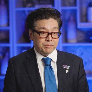 A man in a dark suit, blue tie, and glasses wears a 'GRIFF' pin on his lapel, photographed against a blue-toned background.