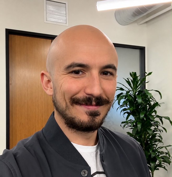 A selfie of a smiling bald man with a beard and mustache, wearing a black jacket over a white shirt. He is in an office setting with a wooden door, a potted plant, and ceiling ductwork visible in the background.