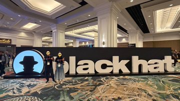 Two people pose in front of a large illuminated "blackhat" sign at what appears to be the Black Hat cybersecurity conference.