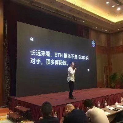 The image shows a man standing at a podium, giving a speech or presentation. The stage has a large screen behind him displaying text written in Chinese characters. There are several people sitting around the room, possibly listening to the speaker or waiting for their turn to speak. A potted plant can be seen near one of the attendees, and there is also a bottle placed on a surface close to the audience. The overall atmosphere suggests that this event might be related to international business or cultural exchange, given the presence of Chinese characters in the speech and the diverse group of people gathered for the presentation.