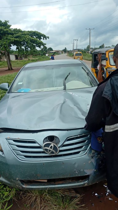 A damaged teal Toyota sedan with a crumpled hood and broken windshield rests on the side of a road, while a person stands beside it and emergency responders gather in the background. The caption references a minor accident with a brother, and the wrecked car in the image illustrates the collision they escaped from unharmed.