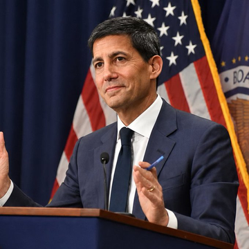 The image features a man wearing a suit and tie standing at a podium with his hands clasped together. He appears to be delivering a speech or addressing an audience, as he is positioned behind a microphone. The backdrop of the scene includes a flag, which could potentially represent the United States, and another flag that might symbolize Bitcoin. The text "If you are under 40, Bitcoin is your new gold." suggests that the man's speech may be related to cryptocurrency or financial matters, possibly discussing the potential benefits or risks associated with Bitcoin for younger generations.

The image also contains a few other objects in the background, such as a book and a cup, which could indicate that this event might be taking place at an office or a similar setting where people gather to discuss various topics.