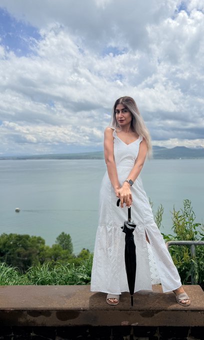 A woman in a flowing white dress poses by a scenic lake, holding a closed black umbrella, under a partly cloudy sky. The image humorously contrasts her poised, elegant demeanor with the post's sarcastic commentary about crypto airdrops and the exaggerated excitement of online hype—suggesting calm detachment amid the financial frenzy.
