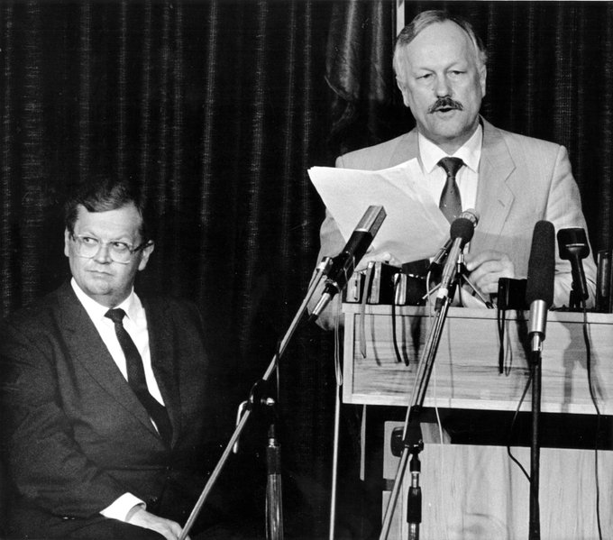 A black and white photograph showing two men in suits; one is seated while the other stands at a podium with multiple microphones, reading from a paper.