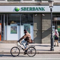 A cyclist rides past a Sberbank branch, with the bank's logo and name clearly visible on the building. The image visually ties to the news of Sberbank, Russia's largest bank, preparing to launch Bitcoin and crypto trading services for its 100+ million clients.
