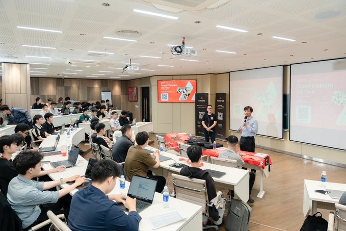 The image shows a classroom full of people sitting at desks, with some individuals using laptops while others are engaged in discussions or listening to a presentation. The room appears to be well-equipped for learning, as there are multiple TVs mounted on the walls displaying information relevant to the coursework being taught. A man is standing near one of these TVs and giving a speech, possibly addressing the class about Avalanche L1s and smart contracts. The overall atmosphere seems to be focused and collaborative, with students actively participating in their education or learning from the speaker.