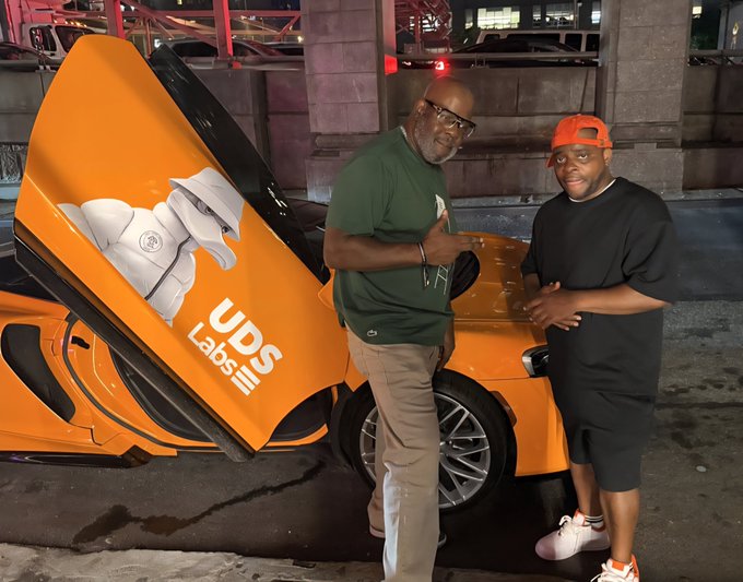 Two men pose next to an orange McLaren supercar with its dihedral door open, which displays the 'UDS LABS' logo featuring an eagle graphic.