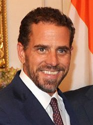 The photo shows a smiling man with dark hair and a goatee, wearing a dark suit and patterned tie. The image accompanies a news update reporting that Hunter Biden states proceeds from the $LAPTOP meme coin will go toward addiction recovery charities, with his team rumored to have made eight figures on the token.