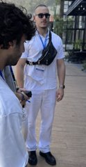 A candid photo of a couple outdoors, with the man in the background wearing a white shirt, black sunglasses, and a small black waist bag, while the woman in the foreground is partially visible.