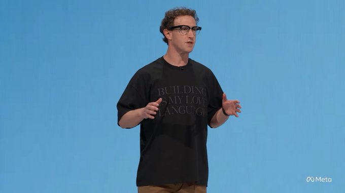 Mark Zuckerberg, wearing a black t-shirt with the phrase "BUILDING IS MY LOVE LANGUAGE," speaks on stage at a Meta event against a blue backdrop, gesturing with his hands as he addresses the audience.