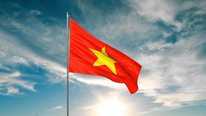 The image shows the red flag of Vietnam with a yellow star waving against a bright blue sky with scattered clouds. This visually represents Vietnam, the country referenced in the post about new regulations imposing fines of up to $1,900 on crypto traders using unlicensed platforms, effective September 1.