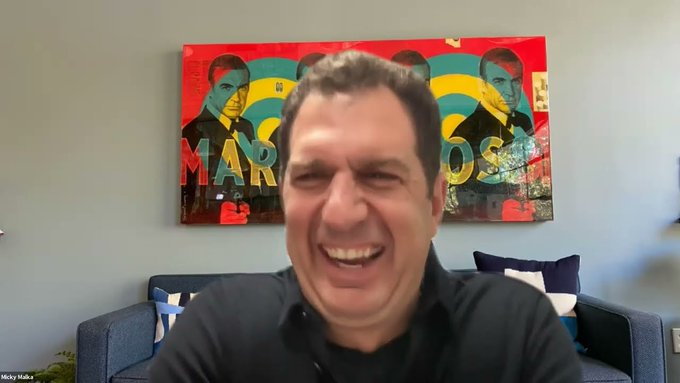 The image shows a man sitting on a couch with his arms crossed, smiling at the camera. The background features a large painting of two men wearing suits, which appears to be a portrait or an abstract art piece. The text "if you hold $HOOD go buy some $TIBBIR thesis" is written in the image, possibly indicating that the man's name is Mick K. O'Malley and he has written a thesis on the subject of $HOOD.