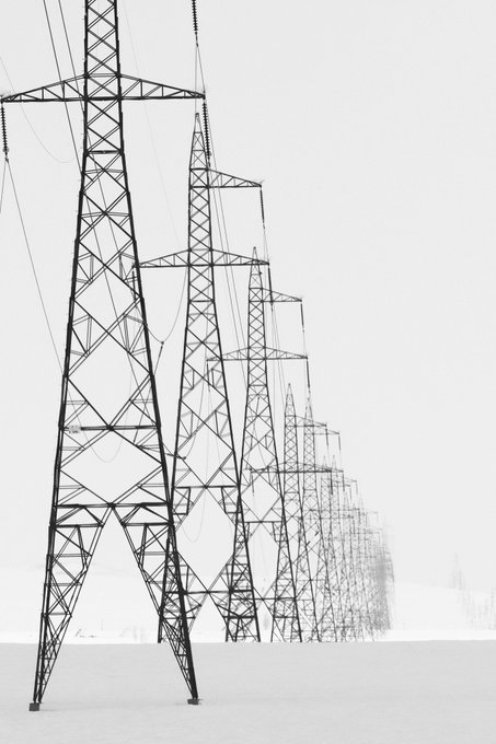A black-and-white photograph showing a row of tall electrical transmission towers stretching in a line into the distance across a snow-covered landscape.