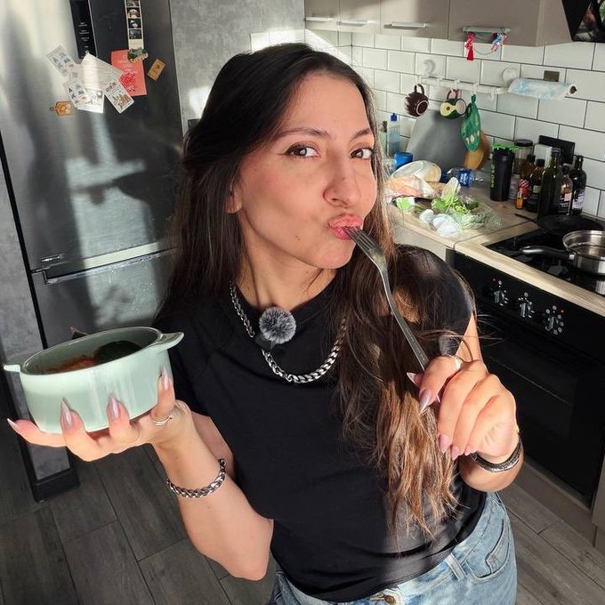 A young woman with long dark hair stands in a kitchen, holding a small green pot of food in one hand while playfully kissing a fork held in the other.