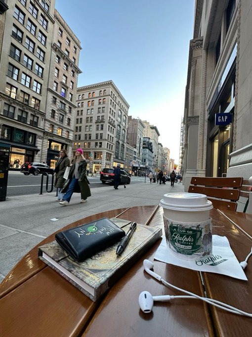 A view from an outdoor café table showing a notebook, pen, coffee cup with 'Ralphs' branding, and earbuds, with a city street scene including a 'GAP' storefront and pedestrians.